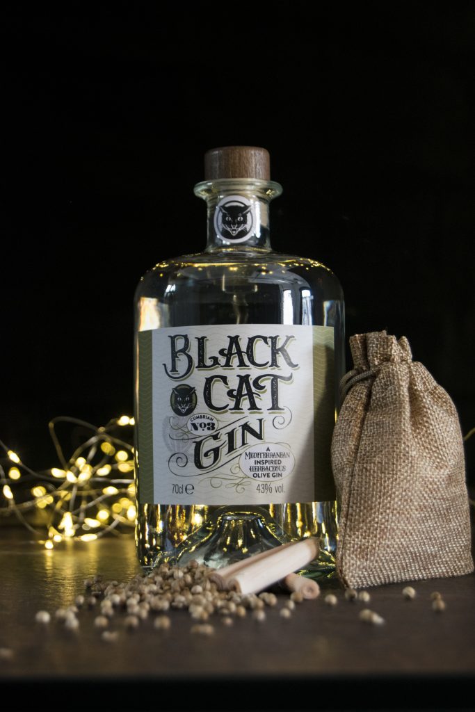 Bottle of Black Cat Savoury Gin Cumbrian No 3 with sparkly lights behind and lemongrass and coriander seeds in the foreground.